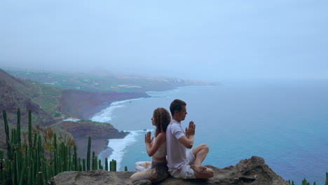 Positioned-on-a-mountain's-rock,-a-man-and-woman-sit-back-to-back,-finding-harmony-through-meditation-and-yoga,-against-the-ocean's-expanse